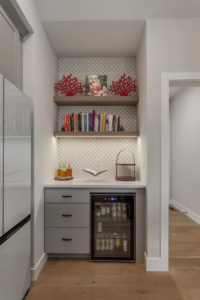 bar with light hardwood / wood-style floors, gray cabinetry, backsplash, stainless steel refrigerator, and wine cooler