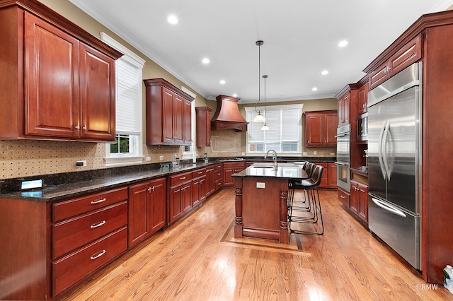 kitchen featuring light hardwood / wood-style floors, tasteful backsplash, built in appliances, an island with sink, and premium range hood