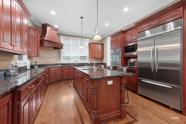 kitchen featuring built in appliances, custom range hood, hardwood / wood-style flooring, and a kitchen island with sink