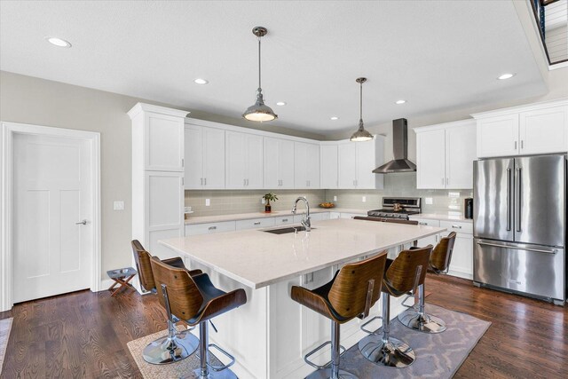 kitchen featuring wall chimney range hood, sink, appliances with stainless steel finishes, a kitchen island with sink, and hanging light fixtures