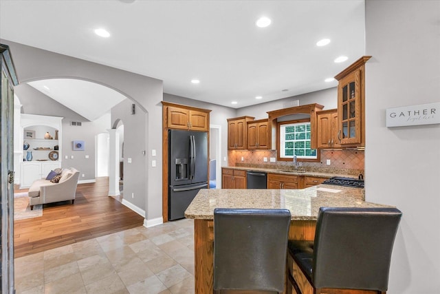 kitchen featuring arched walkways, brown cabinetry, light stone counters, black appliances, and a sink