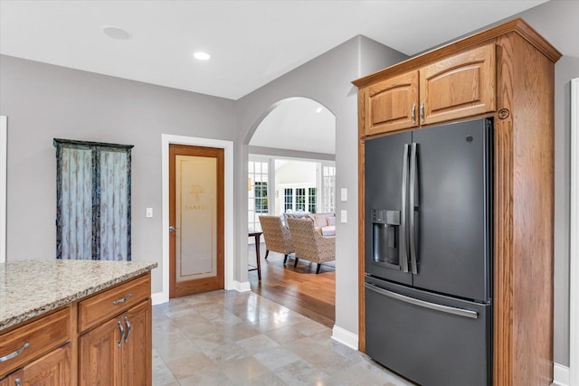 kitchen featuring baseboards, arched walkways, brown cabinets, light stone countertops, and fridge with ice dispenser