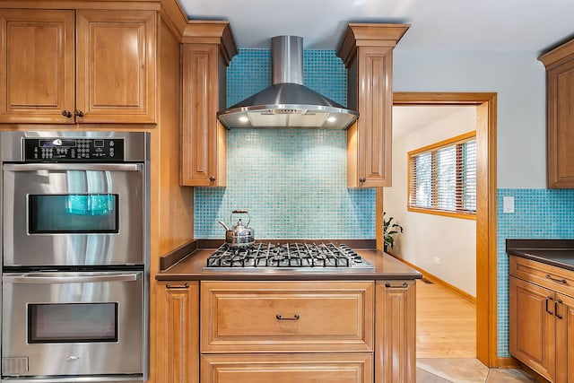 kitchen with wall chimney exhaust hood, backsplash, stainless steel appliances, dark countertops, and brown cabinetry