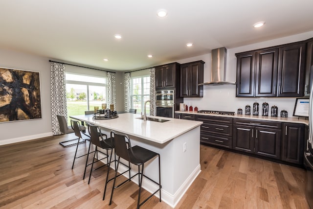 kitchen featuring a center island with sink, stainless steel appliances, light wood-type flooring, wall chimney range hood, and sink