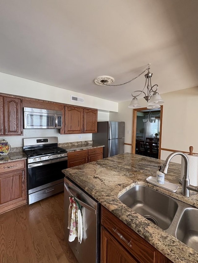 kitchen with appliances with stainless steel finishes, hanging light fixtures, dark wood-type flooring, sink, and dark stone countertops