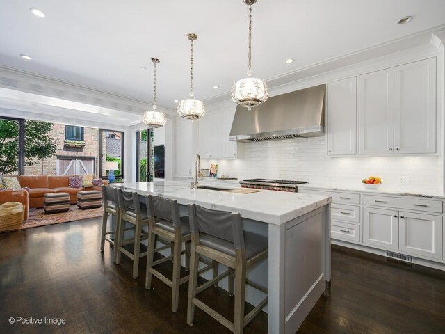 kitchen with wall chimney range hood, white cabinetry, dark wood-type flooring, a center island with sink, and backsplash