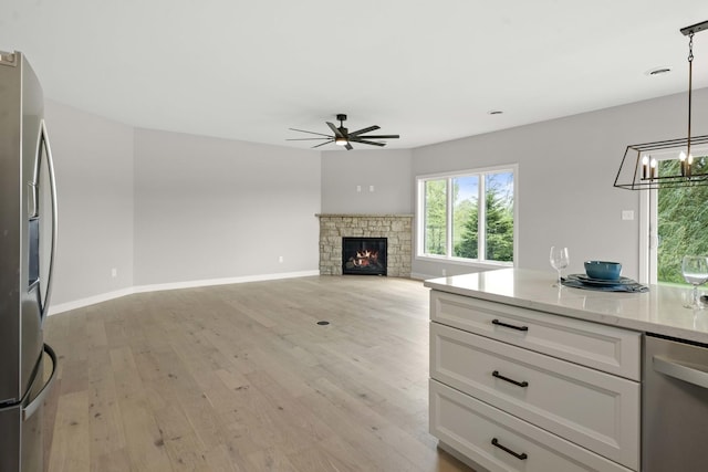 unfurnished living room with baseboards, a stone fireplace, light wood finished floors, and ceiling fan with notable chandelier