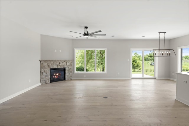 unfurnished living room featuring baseboards, light wood finished floors, ceiling fan with notable chandelier, and a stone fireplace
