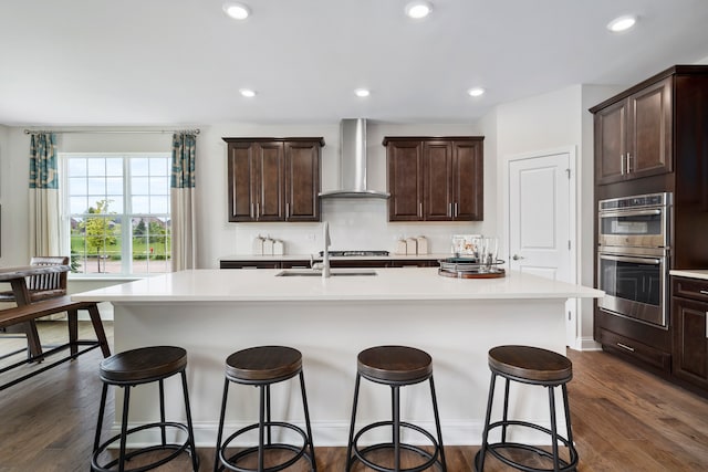 kitchen with wall chimney range hood, dark wood-type flooring, double oven, and a center island with sink