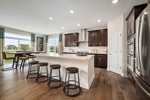 kitchen with wall chimney range hood, dark wood-type flooring, a center island with sink, and stainless steel appliances