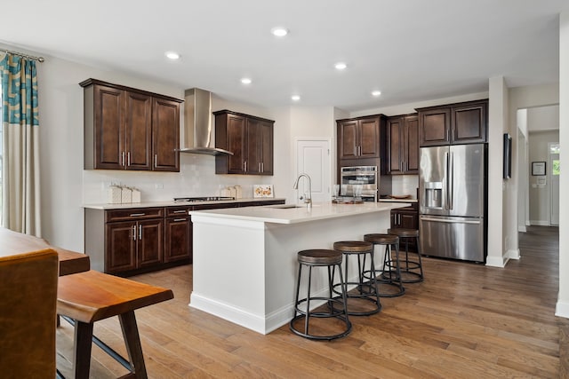 kitchen with wall chimney range hood, stainless steel fridge with ice dispenser, a center island with sink, light hardwood / wood-style flooring, and tasteful backsplash