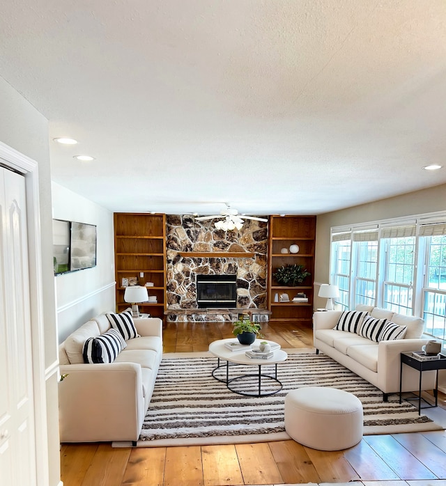 living area with wood finished floors, a textured ceiling, a stone fireplace, built in shelves, and recessed lighting