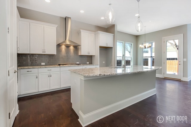 kitchen featuring hanging light fixtures, wall chimney exhaust hood, light stone countertops, an island with sink, and white cabinetry