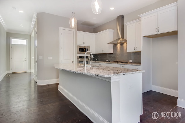 kitchen featuring light stone counters, stainless steel appliances, wall chimney range hood, white cabinetry, and an island with sink