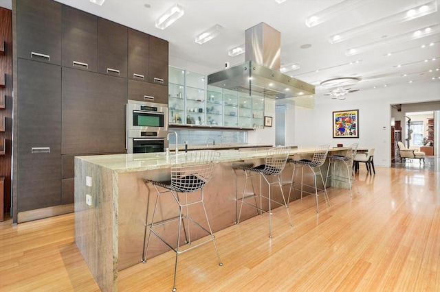 kitchen featuring a large island, light stone counters, light hardwood / wood-style flooring, island exhaust hood, and a breakfast bar area