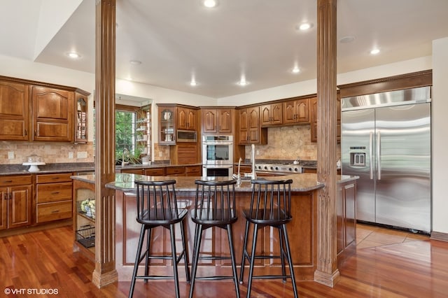 kitchen with appliances with stainless steel finishes, hardwood / wood-style floors, an island with sink, and dark stone counters