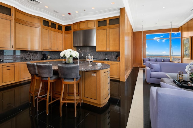 kitchen with dark stone counters, an island with sink, sink, hanging light fixtures, and ornamental molding