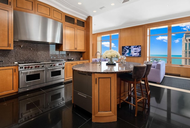 kitchen with range with two ovens, an island with sink, decorative backsplash, ornamental molding, and a water view