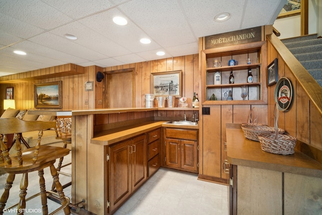 bar featuring light tile patterned flooring, sink, wood walls, and a drop ceiling