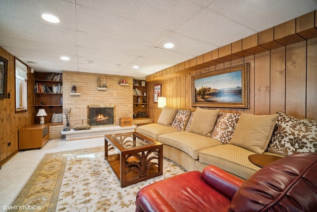 living room with a drop ceiling, wooden walls, built in features, a brick fireplace, and brick wall