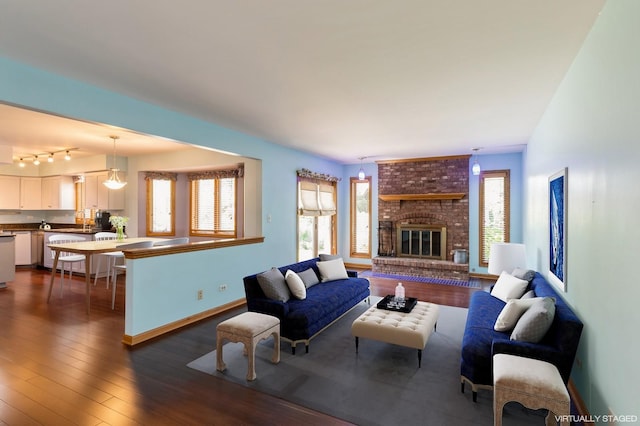 living room featuring track lighting, dark wood-style flooring, a brick fireplace, and baseboards