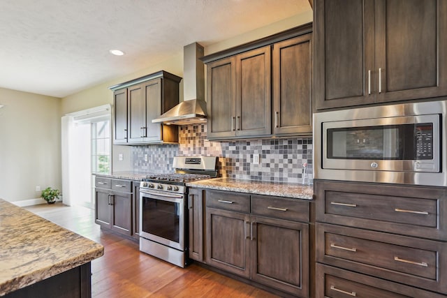 kitchen featuring tasteful backsplash, dark brown cabinetry, wood finished floors, stainless steel appliances, and wall chimney exhaust hood