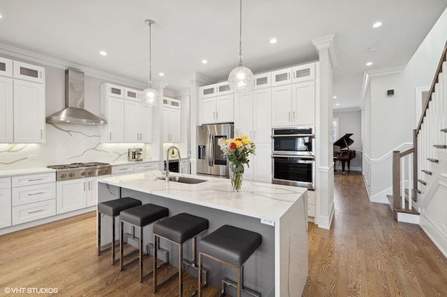 kitchen featuring white cabinetry, sink, stainless steel appliances, wall chimney range hood, and a center island with sink