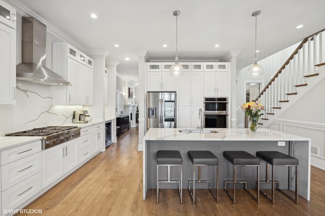 kitchen with stainless steel appliances, a spacious island, sink, wall chimney range hood, and white cabinetry