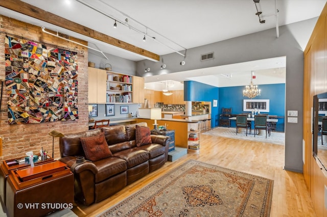 living room with brick wall, light wood-type flooring, a notable chandelier, track lighting, and beam ceiling