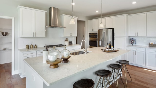 kitchen featuring wall chimney range hood, hanging light fixtures, an island with sink, sink, and appliances with stainless steel finishes