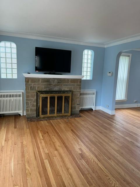 unfurnished living room with wood finished floors, radiator, a stone fireplace, and ornamental molding