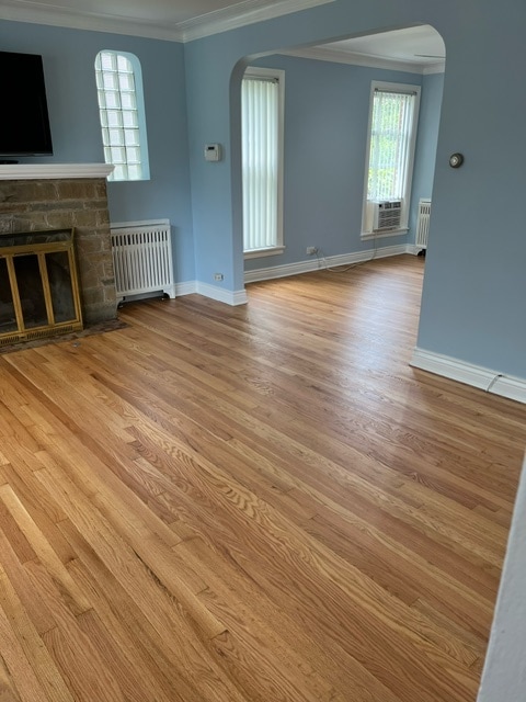 unfurnished living room featuring wood-type flooring, crown molding, cooling unit, radiator, and a stone fireplace