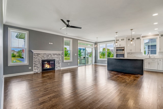 unfurnished living room featuring ceiling fan, ornamental molding, sink, a stone fireplace, and dark wood-type flooring