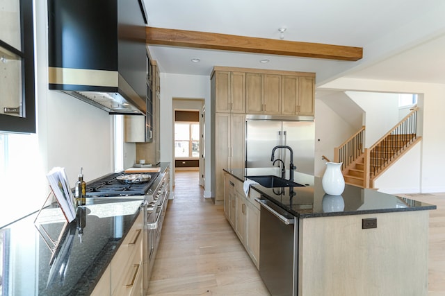 kitchen with dark stone counters, wall chimney range hood, a center island with sink, high end appliances, and light hardwood / wood-style flooring