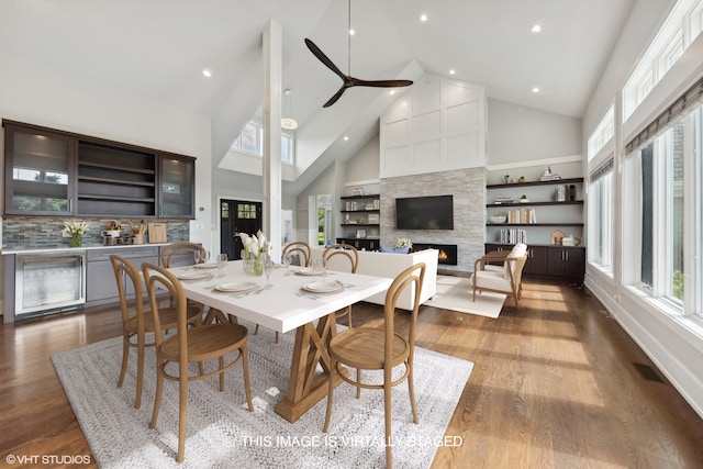 dining area featuring ceiling fan, beverage cooler, dark hardwood / wood-style flooring, a large fireplace, and high vaulted ceiling