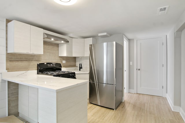 kitchen featuring stainless steel fridge, light stone counters, kitchen peninsula, black gas range, and wall chimney exhaust hood