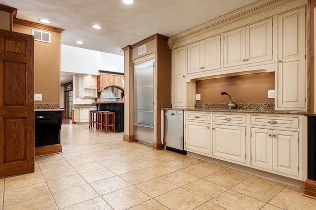 kitchen with dishwashing machine, light tile patterned floors, cream cabinetry, stone counters, and sink