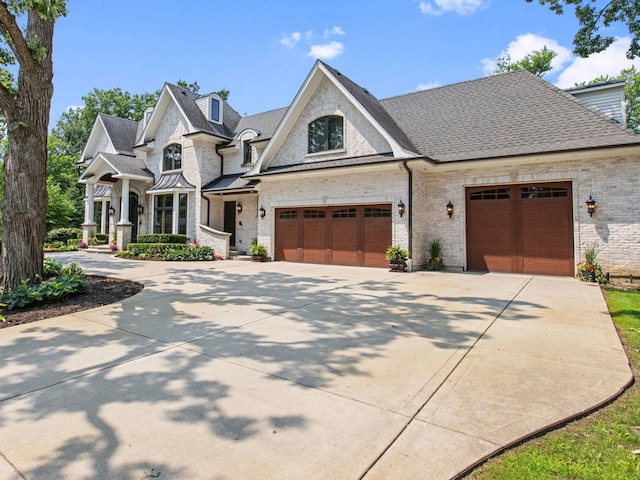 french country inspired facade with a garage, driveway, brick siding, and a shingled roof