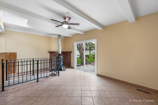 tiled spare room featuring beamed ceiling, a wood stove, and ceiling fan