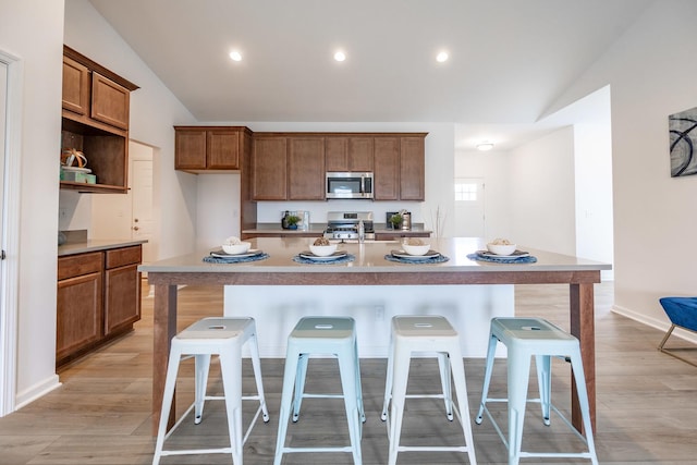 kitchen featuring a kitchen island with sink, stainless steel appliances, light countertops, brown cabinetry, and a kitchen bar