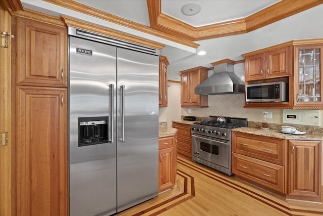 kitchen with high end appliances, decorative backsplash, brown cabinets, crown molding, and wall chimney range hood