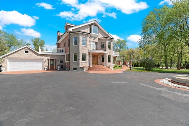 view of front facade with an attached garage, a balcony, stone siding, driveway, and a chimney