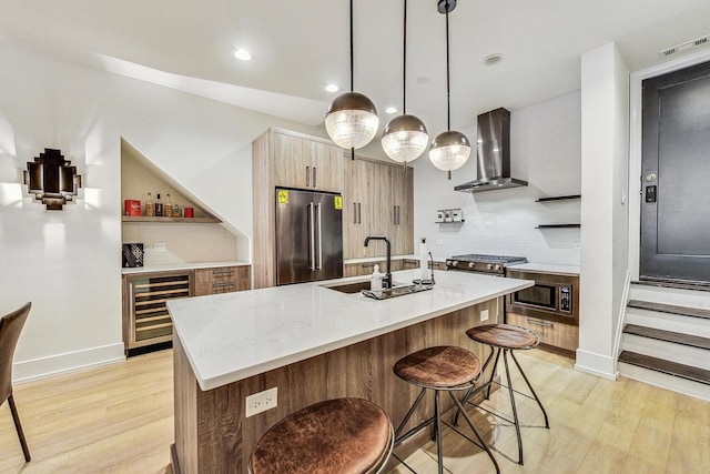 kitchen with stainless steel appliances, beverage cooler, wall chimney range hood, pendant lighting, and light hardwood / wood-style floors