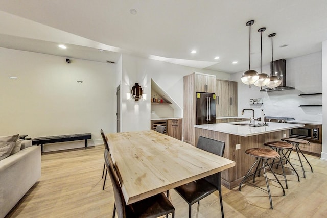 kitchen featuring light wood-type flooring, wall chimney exhaust hood, a kitchen island with sink, sink, and hanging light fixtures