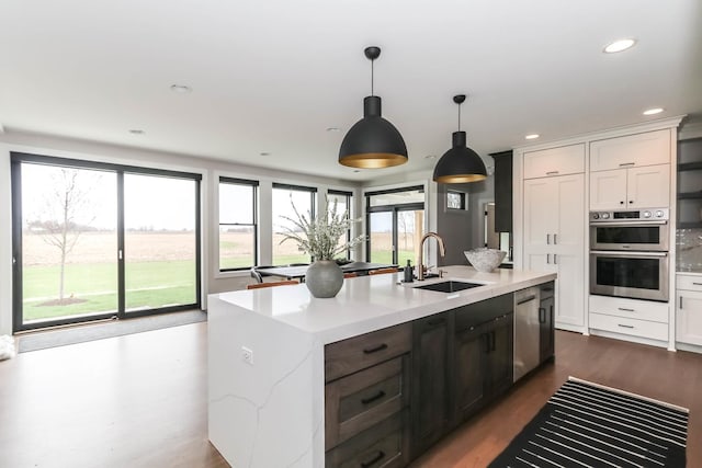 kitchen with a kitchen island with sink, stainless steel appliances, a sink, white cabinetry, and decorative light fixtures