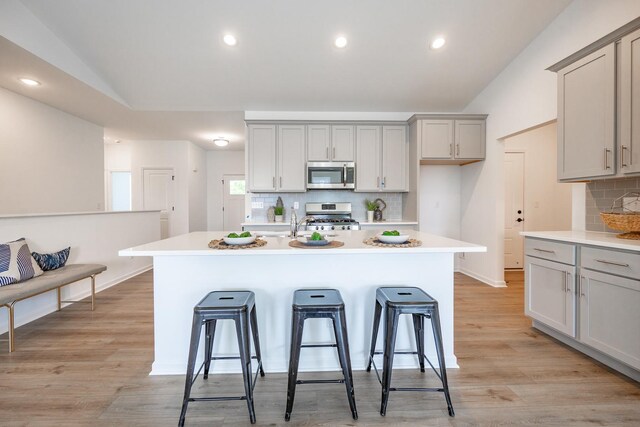 kitchen featuring tasteful backsplash, an island with sink, light hardwood / wood-style floors, vaulted ceiling, and stove