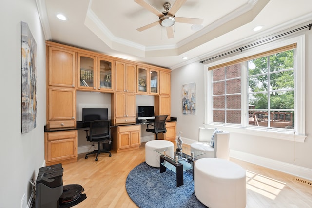 office area with light wood-type flooring, a raised ceiling, built in desk, ornamental molding, and ceiling fan