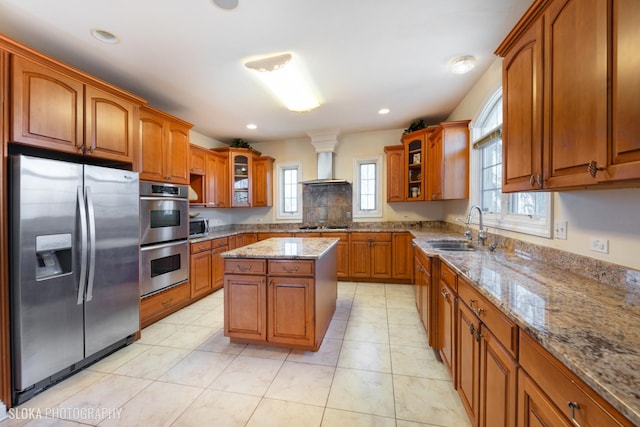 kitchen featuring sink, wall chimney range hood, stainless steel appliances, light stone counters, and a kitchen island