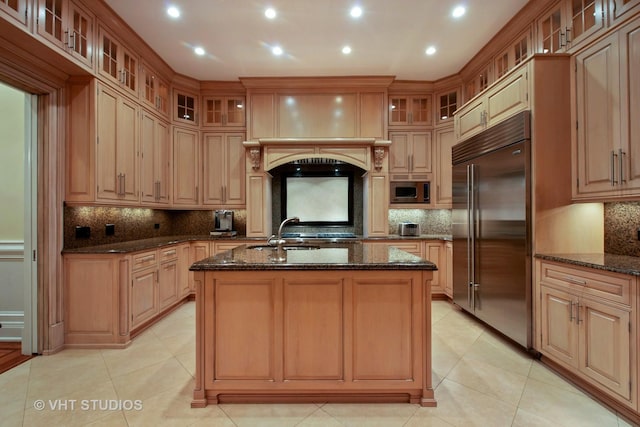 kitchen featuring built in appliances, dark stone counters, a sink, and a kitchen island with sink