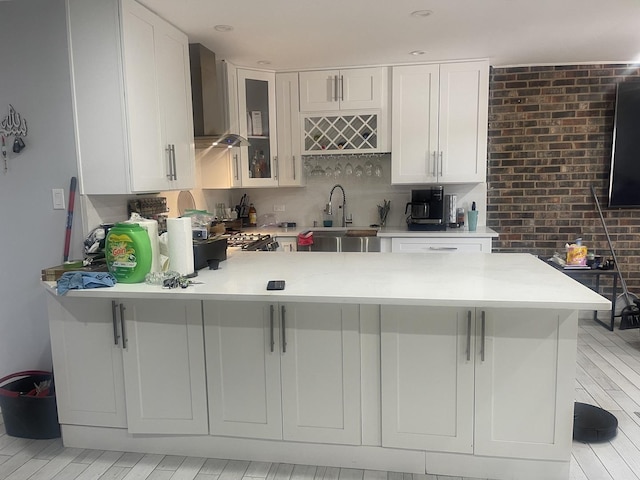 kitchen featuring wall chimney range hood, a peninsula, light countertops, a sink, and glass insert cabinets
